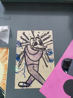 Close-up of a sticker featuring a cartoon-like figure walking, seemingly with needles or sticks protruding from its head. The art style is expressive and slightly rough around the edges, with visible brushstrokes or printmaking techniques. The color palette consists mainly of purple, blue, and beige. The sticker is adhered to a dark gray surface.