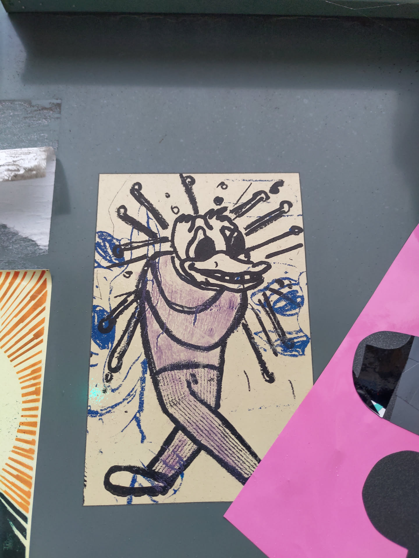 Close-up of a sticker featuring a cartoon-like figure walking, seemingly with needles or sticks protruding from its head. The art style is expressive and slightly rough around the edges, with visible brushstrokes or printmaking techniques. The color palette consists mainly of purple, blue, and beige. The sticker is adhered to a dark gray surface.