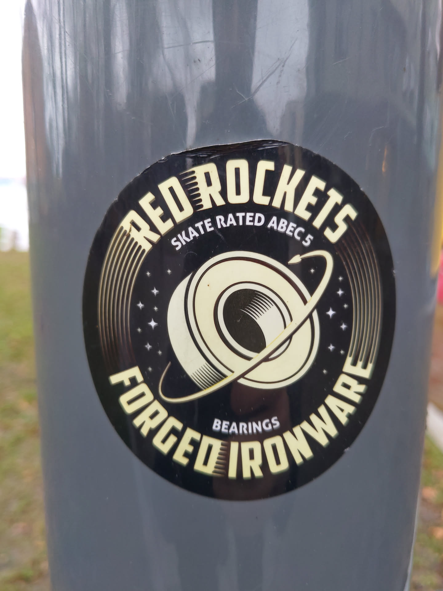 A round sticker with a space theme, featuring a stylized bearing in the center. The text 'Red Rockets' is at the top, 'Forged Ironware' at the bottom, with 'Skate Rated ABEC 5' in between. The design is primarily dark with gold/yellow accents.