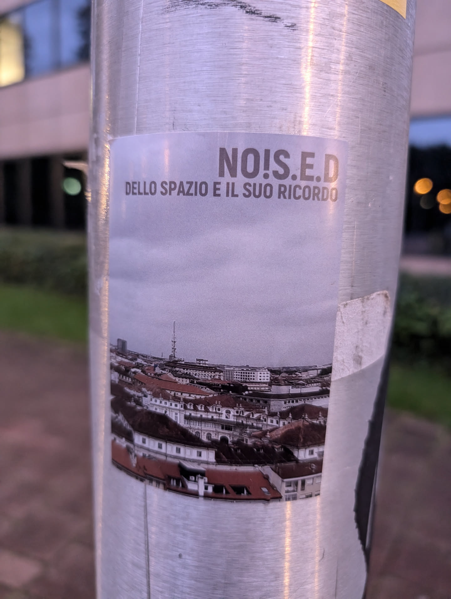 A sticker featuring the text "NO!S.E.D" and the Italian phrase "DELLO SPAZIO E IL SUO RICORDO" which translates to "OF SPACE AND ITS MEMORY", along with an image of an aerial view of a city, possibly in Europe.  The sticker is affixed to a metal pole.