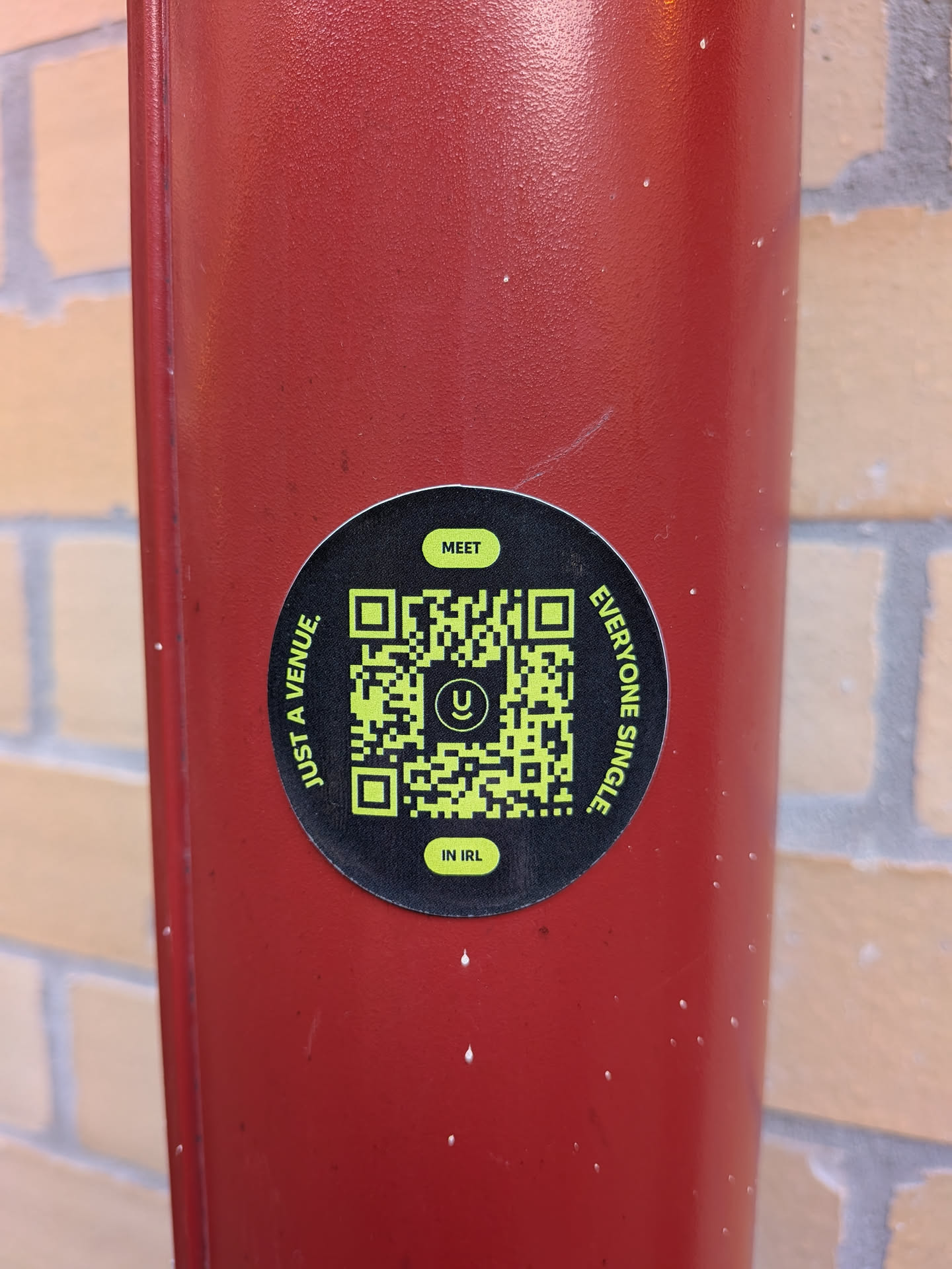 A circular black sticker with a bright green QR code at its center. The QR code has a small, stylized 'U' logo within it. Text is arranged around the QR code in a circular fashion. At the top, in a small green oval, is the word 'MEET'. Along the left side of the circle, in green text, reads 'JUST A VENUE.'. Along the right side, also in green text, reads 'EVERYONE SINGLE.'. At the bottom, in another small green oval, is the text 'IN IRL.'. The sticker is affixed to a textured red surface.