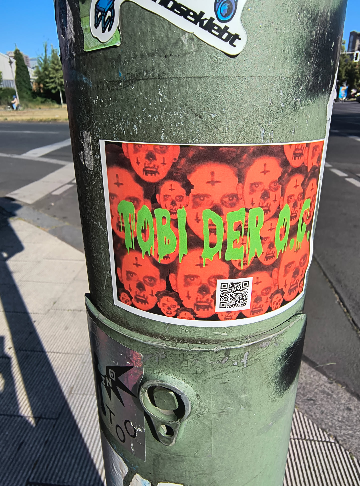 The sticker is a rectangular print with a white border, affixed to a weathered green pole. Its central image is a dark, reddish-orange background filled with a dense, repeating pattern of unsettling faces. These faces resemble skulls or zombies, featuring prominent eye sockets and mouths, and many are marked with small, black inverted crosses on their foreheads. Superimposed over this macabre collage is the text 'TOBI DER O.G.' rendered in a vibrant, slime-green font that appears to be dripping. A small, black and white QR code is visible in the bottom right corner of the sticker.