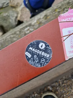 A round black and white sticker with illustrations and text.  The text says "Magdebier" prominently, with smaller text that reads "Das Bier für Magdeburg" (The beer for Magdeburg) and "Lokalo".  The illustrations depict people and buildings, suggesting a local brewery or beer brand from Magdeburg.