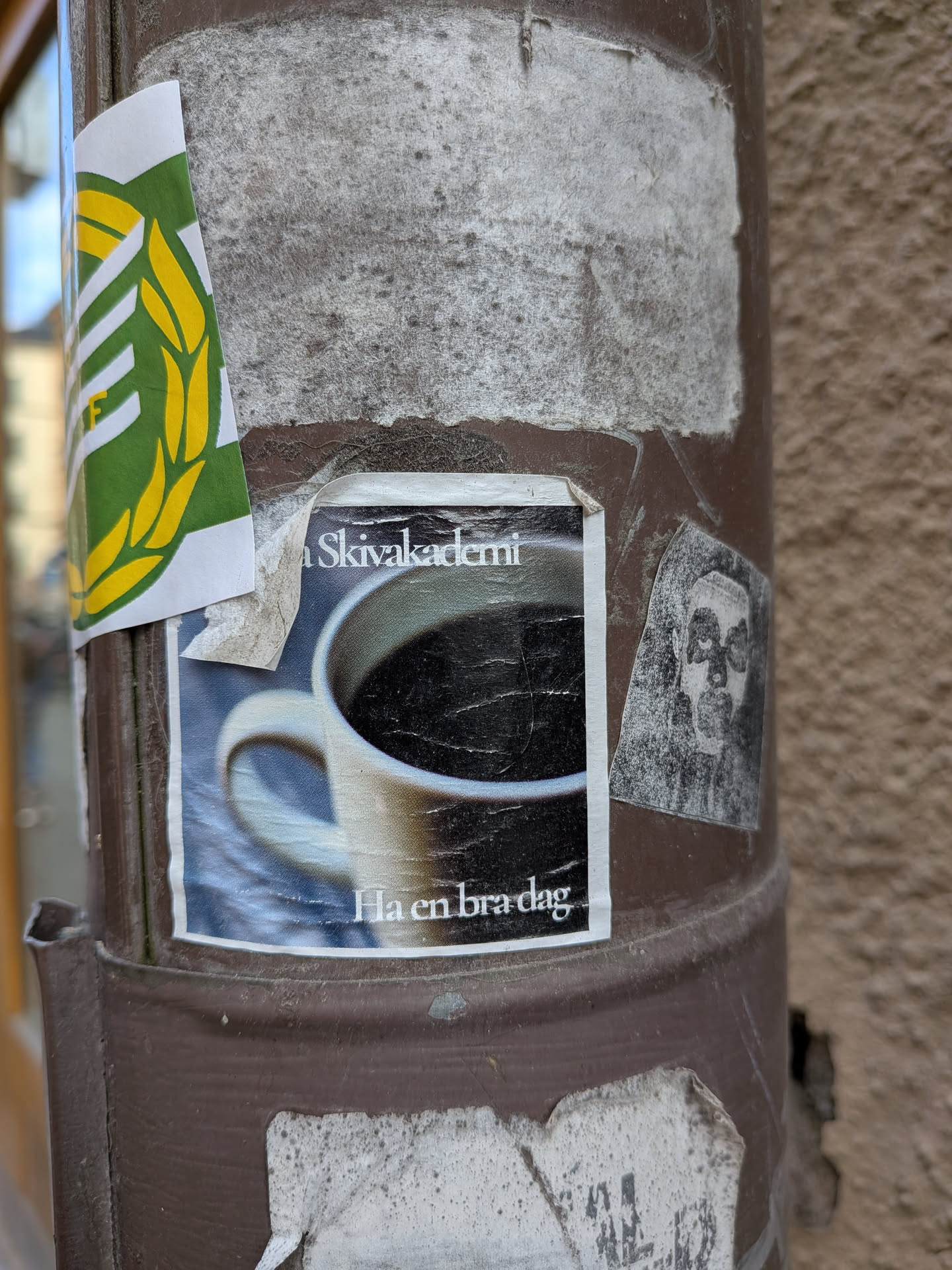 The central sticker features a close-up, slightly angled photograph of a white or light-colored ceramic coffee mug filled with dark, black coffee. The coffee surface reflects some light. The mug's handle is visible on the left side. The background is a blurred gradient of blue and grey tones. White text is present above and below the mug. The sticker itself is rectangular, shows signs of wear with creases and a peeling top-right corner, and is adhered to a brown pole.