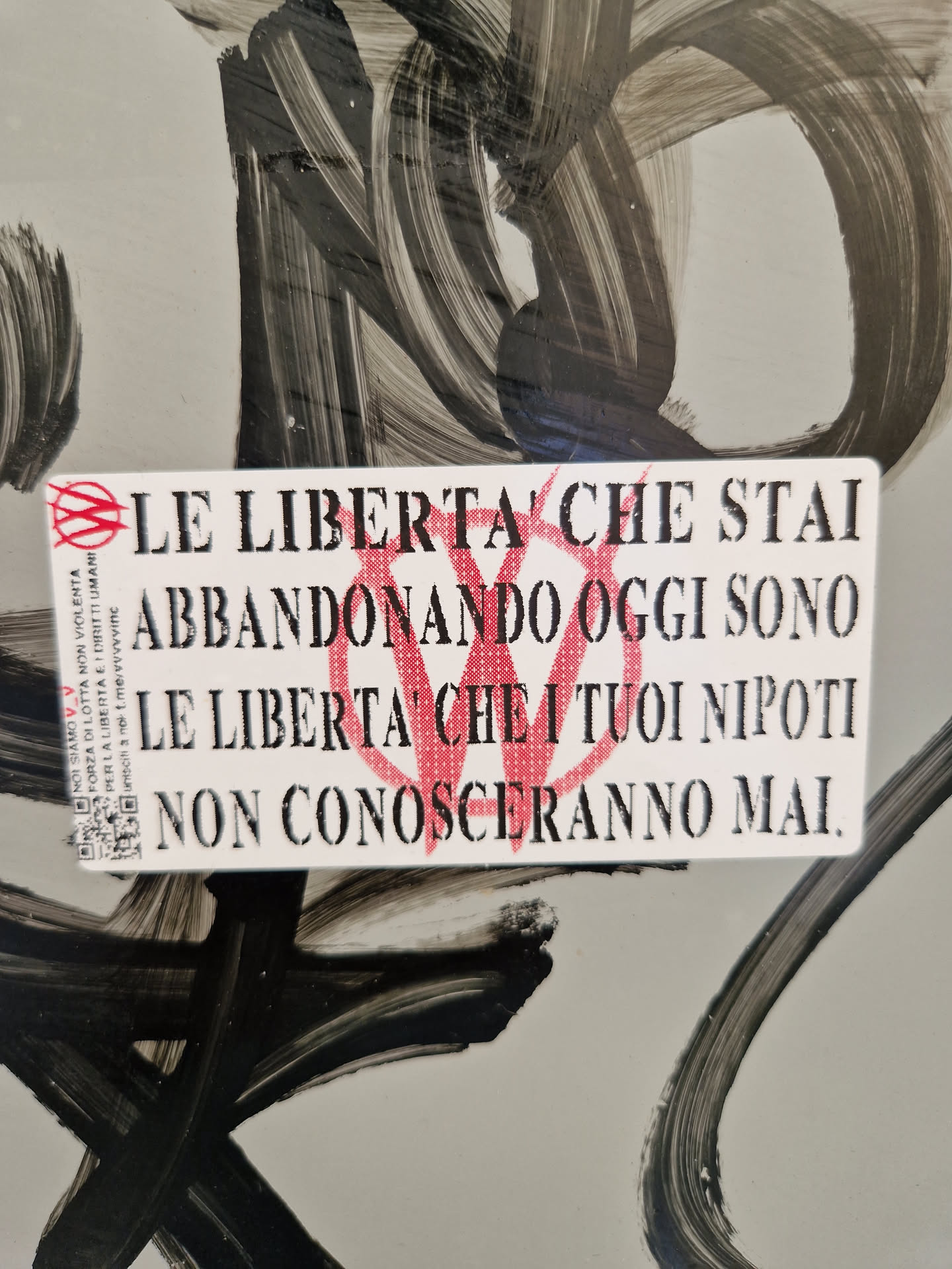 A sticker with a quote in Italian about lost freedoms. The quote is printed in black ink on a white background, with a red symbol at the beginning and end of the text. A QR code is located at the bottom-left corner. The sticker is placed on a surface with black paint strokes.