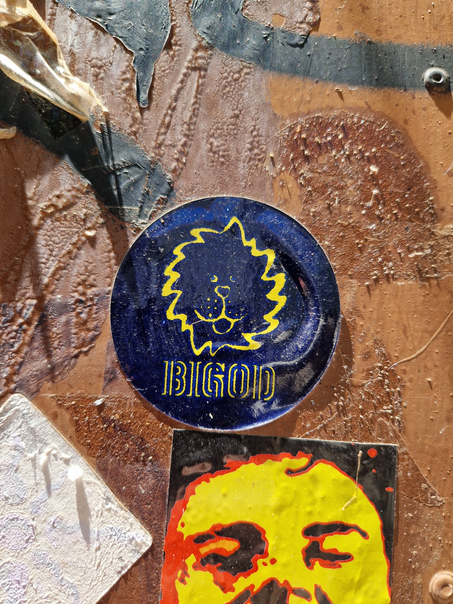 Round blue sticker with a yellow lion-like creature and the word BIGOD written below it. The sticker is affixed to a textured surface with other stickers around it.