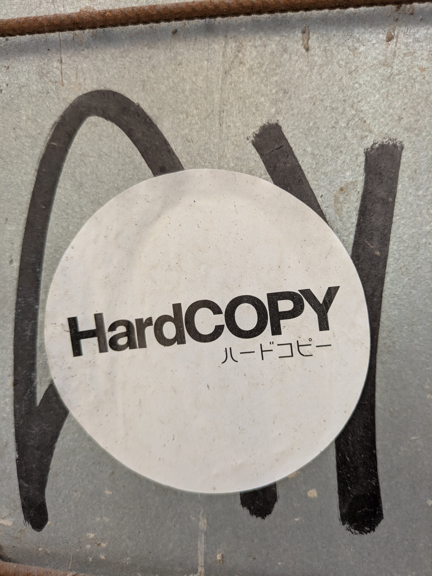 The image shows a round, white sticker with the text 'HardCOPY' in bold, black letters. Below that, there is Japanese text that also translates to 'Hard Copy'. The sticker is attached to a corrugated metal surface which is also covered with black graffiti.