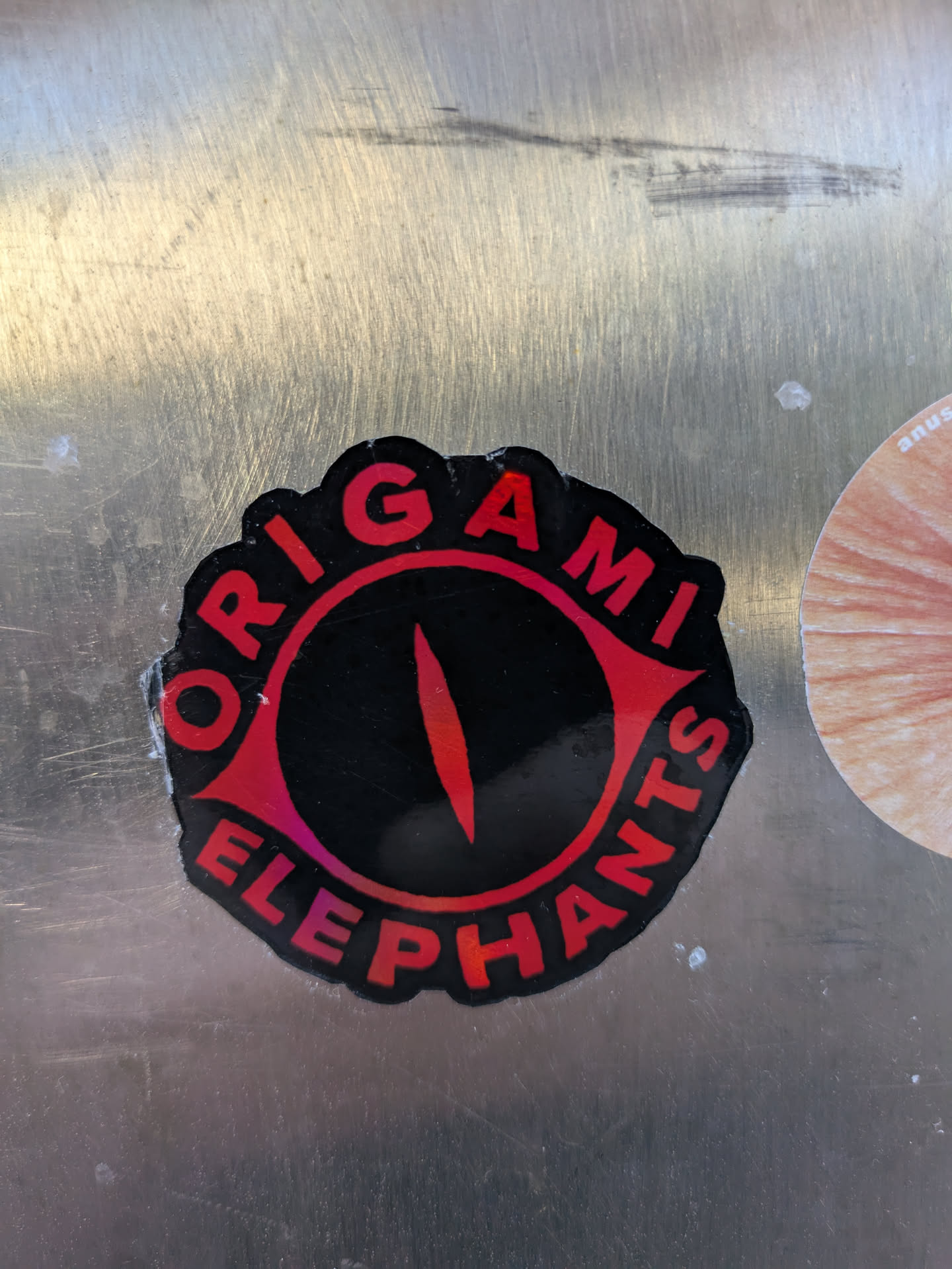 This sticker features a striking red and black eye design, with the text 'ORIGAMI ELEPHANTS' encircling it. The eye has a black center and a red iris. The text is in red and bold. The background surface is a brushed metal.