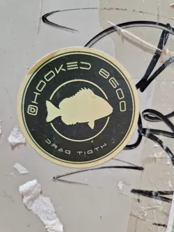 Round sticker with a fish illustration and text. The text includes "HOOKED 8600" and "DRAG TIGHT". There is also an instagram logo.