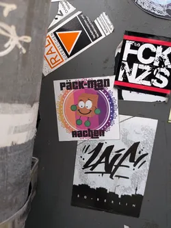 Street sticker A square white sticker featuring a smiling brown cardboard parcel character with cartoon eyes and round green hands and feet. The character is placed inside an ornate circular mandala-style frame with a warm orange-to-purple gradient background. Bold, stylized black lettering reads 'päck-man' above the circle and 'aachen' below it.