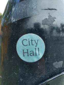 A teal round sticker with the words "City Hall" in a simple sans-serif font.  It's affixed to a dark-colored surface that appears to be outdoors.