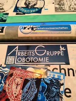 A rectangular sticker with a dark blue background and white text and graphics. The main text reads 'ARBEITSGRUPPE LOBOTOMIE' (Working Group Lobotomy). The 'A' in 'ARBEITS' and the 'G' in 'GRUPPE' are stylized with prominent, angular designs. A small white dove, containing a symbol resembling a peace sign, is depicted flying above the 'R' in 'GRUPPE'. A functional QR code is located in the bottom left corner of the sticker. The entire design is framed by a thin white border.