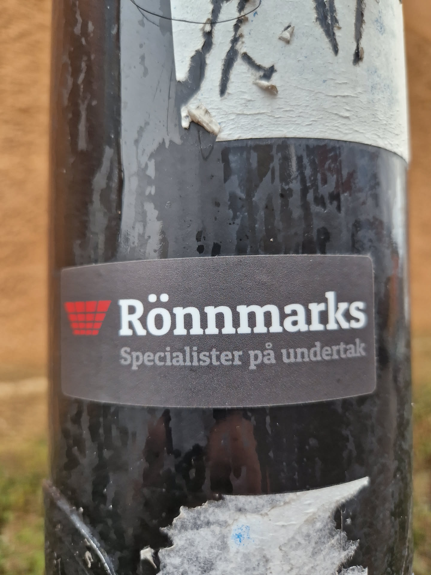 A black and red sticker for Rönnmarks, specialists in undertaks. The sticker is attached to a dark-colored pole outdoors.