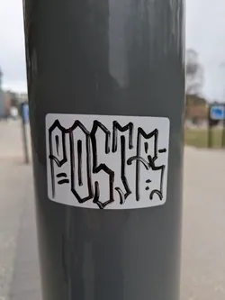 The image shows a white sticker adhered to a gray pole. The sticker features the word "POSITIF" written in a stylized, graffiti-inspired font. The letters are thick and black, outlined to create a bold, almost 3D effect. The background of the sticker is white, and it has rounded corners.