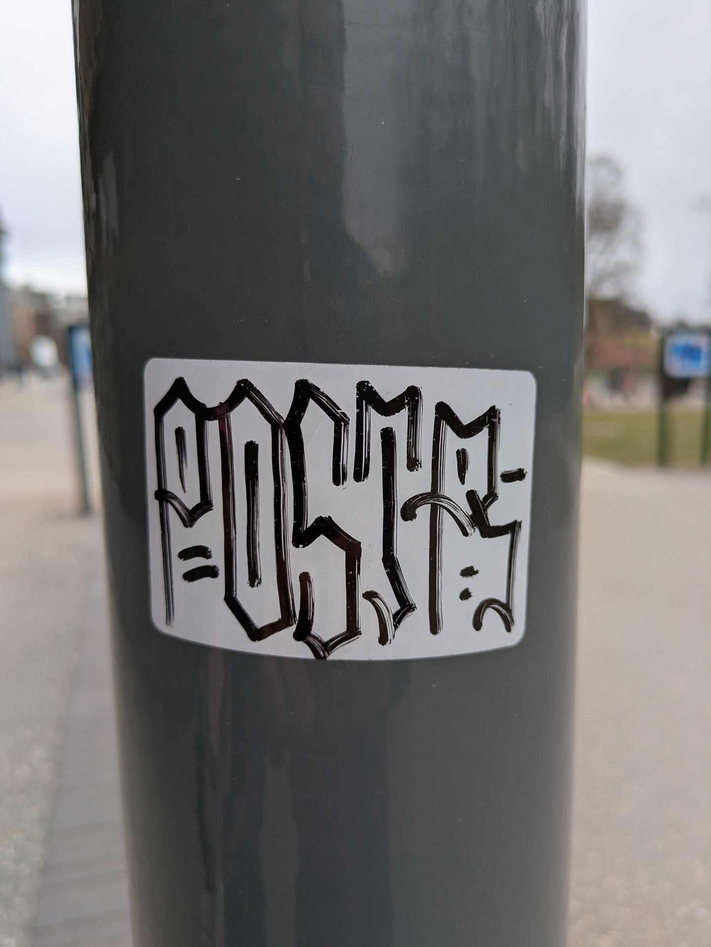 The image shows a white sticker adhered to a gray pole. The sticker features the word "POSITIF" written in a stylized, graffiti-inspired font. The letters are thick and black, outlined to create a bold, almost 3D effect. The background of the sticker is white, and it has rounded corners.