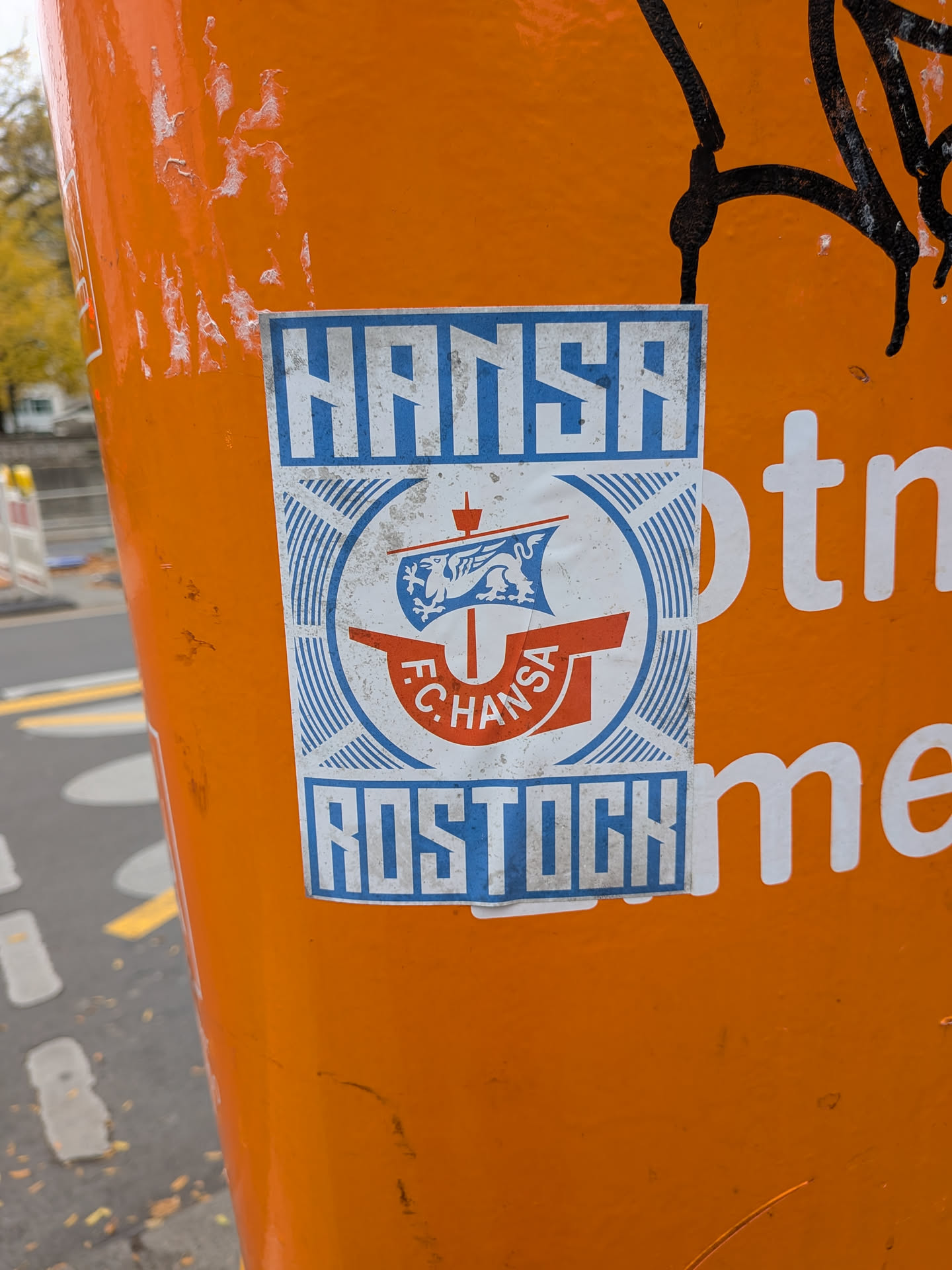 A rectangular sticker adhered to a bright orange surface, featuring the iconography of German football club F.C. Hansa Rostock. At the top, 'HANSA' is written in stylized, blocky white font on a blue rectangular banner. At the bottom, 'ROSTOCK' is similarly stylized in blue letters on a white field bordered by blue. In the center is the club's circular crest, depicting a Hanseatic cog ship in red and white with a blue sail bearing a white griffin, and the text 'F.C. HANSA' along the hull, set against a backdrop of radiating blue and white concentric and linear patterns.
