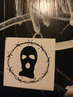 A black and white sticker of a balaclava surrounded by barbed wire. The style is minimalist and edgy.