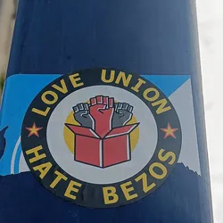 A sticker expressing support for unions and opposition to Jeff Bezos.  It features three raised fists emerging from a box, symbolizing unity and action. The circular design incorporates the text "Love Union Hate Bezos" with stars on either side.