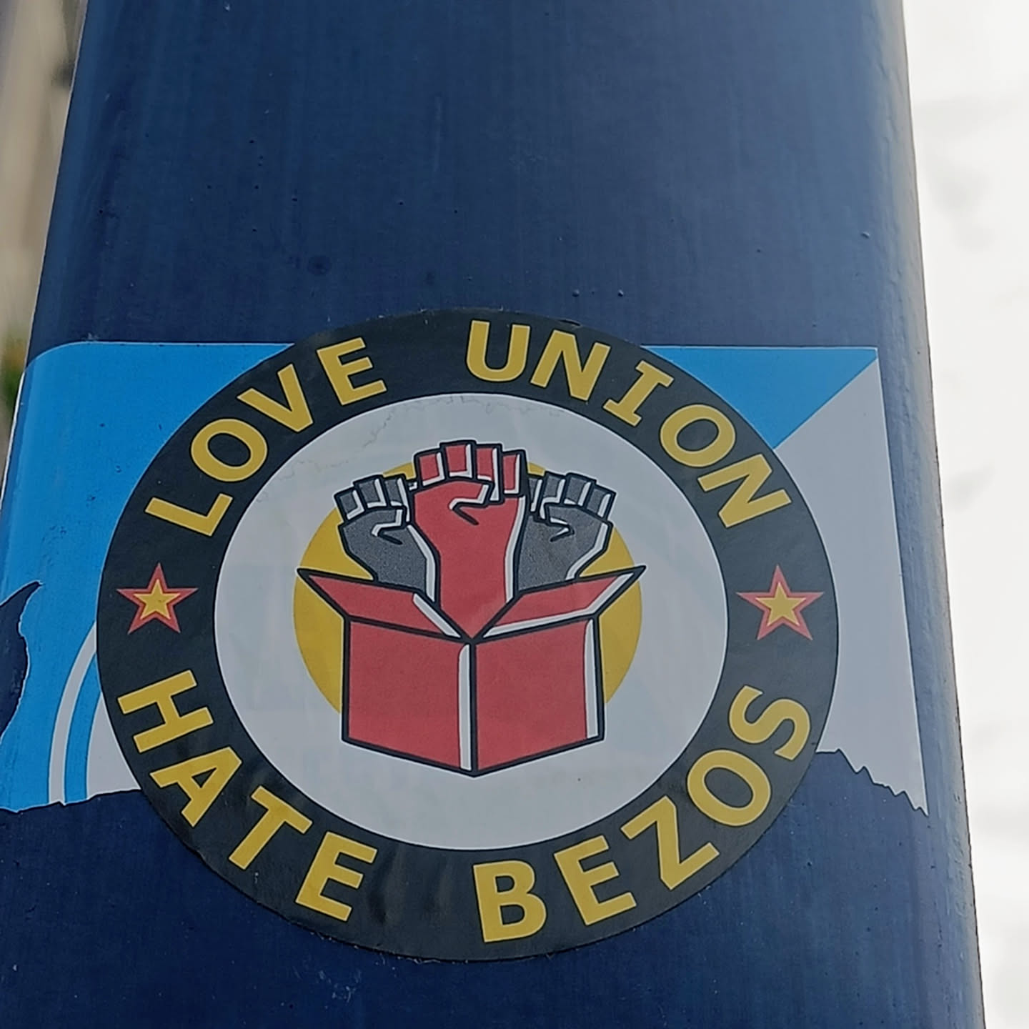 A sticker expressing support for unions and opposition to Jeff Bezos.  It features three raised fists emerging from a box, symbolizing unity and action. The circular design incorporates the text "Love Union Hate Bezos" with stars on either side.