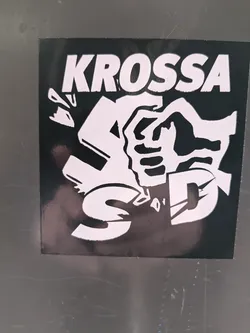 A black and white sticker with a bold graphic design. The central motif is two hands clasped, depicted in a way that suggests both unity and potential separation. The word KROSSA is written in a large, sans-serif font at the top. The style is reminiscent of protest art or political graphics. 