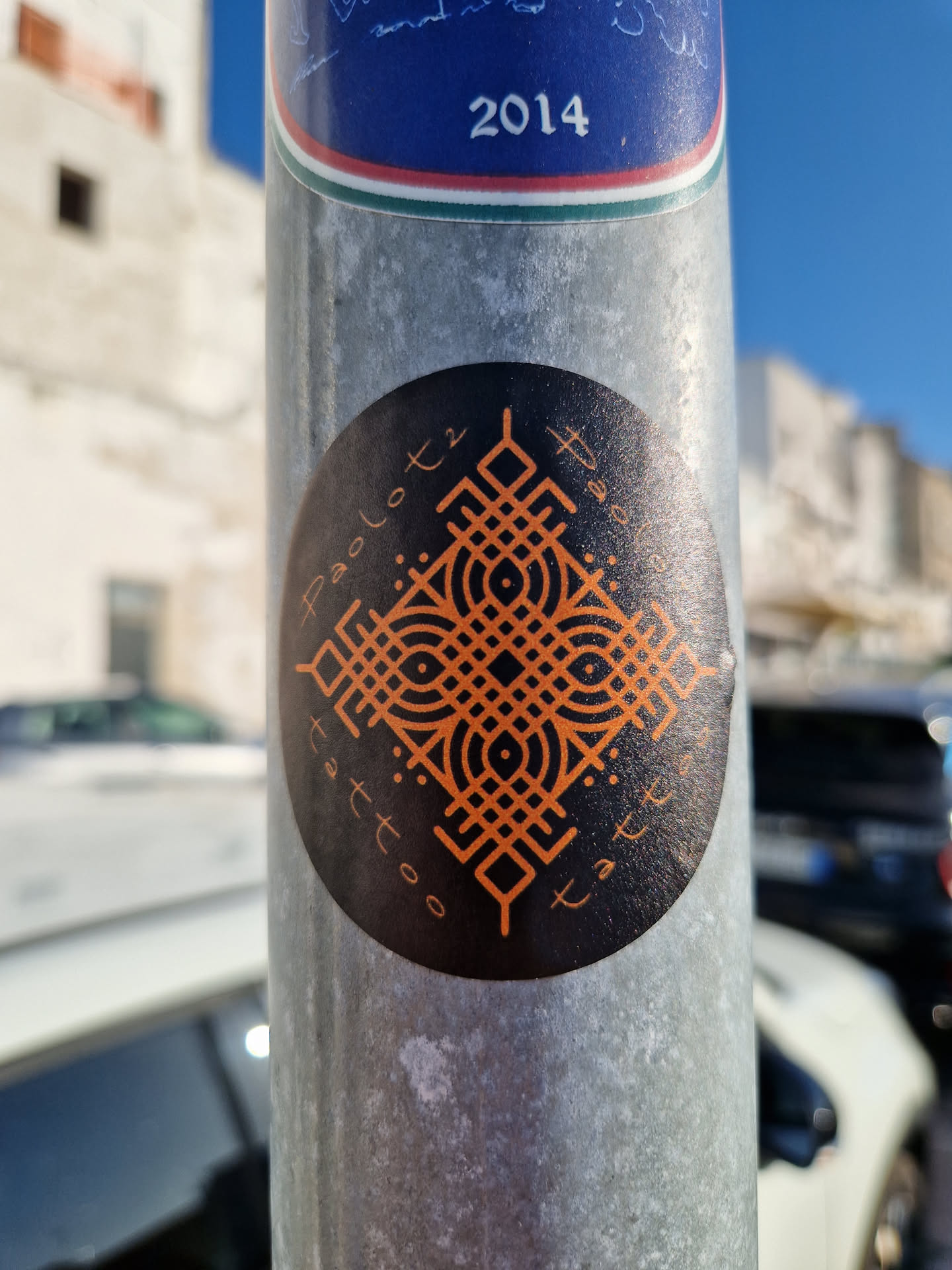 Round black and orange sticker with geometric pattern and the text "Apollo Tattoo" written in a circular style around the central design. The style is reminiscent of traditional tattoo designs.