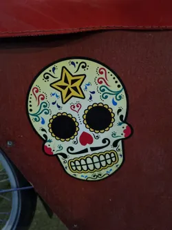 A vibrant sticker depicting a sugar skull, a traditional symbol of Dia de los Muertos (Day of the Dead). The skull is adorned with intricate floral designs and a star. It is affixed to a reddish-brown surface.