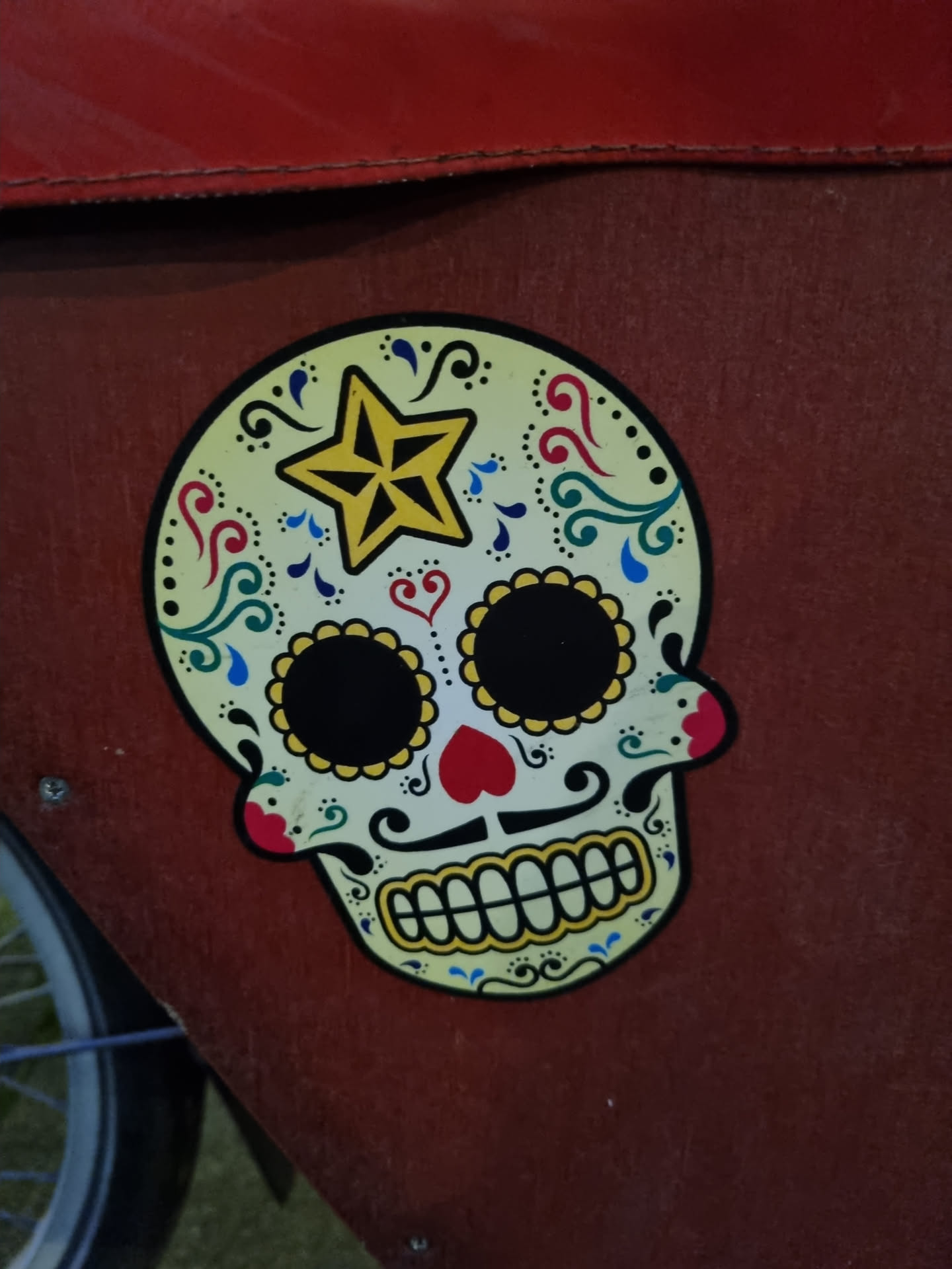 A vibrant sticker depicting a sugar skull, a traditional symbol of Dia de los Muertos (Day of the Dead). The skull is adorned with intricate floral designs and a star. It is affixed to a reddish-brown surface.