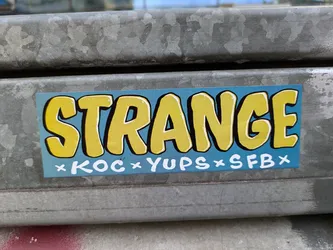 A rectangular sticker with a light blue background and the word "STRANGE" in bold, yellow, outlined letters. Smaller white text underneath reads "×KOC×YUPS×SFB×". The sticker is affixed to a weathered metal surface.