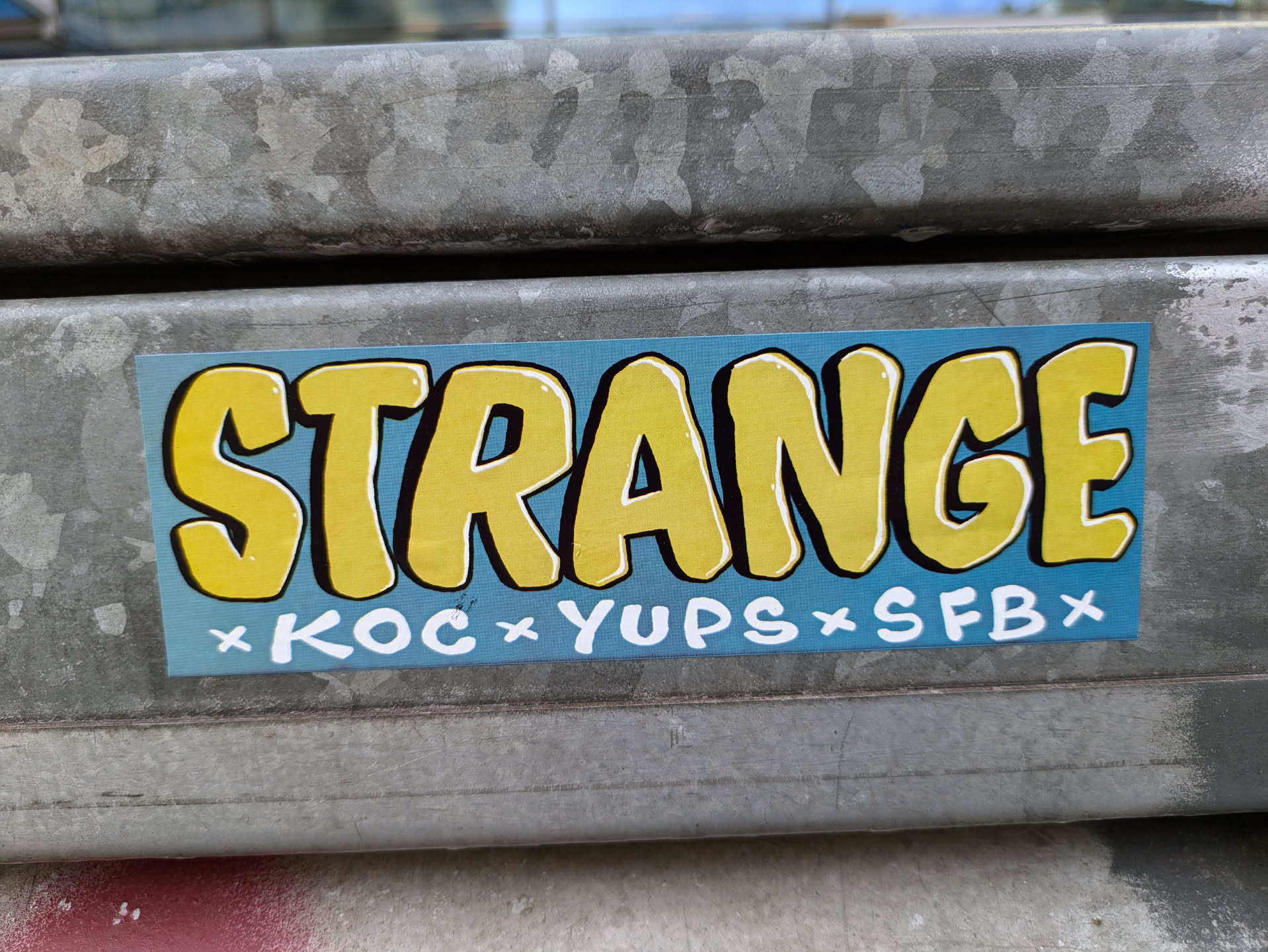 A rectangular sticker with a light blue background and the word "STRANGE" in bold, yellow, outlined letters. Smaller white text underneath reads "×KOC×YUPS×SFB×". The sticker is affixed to a weathered metal surface.