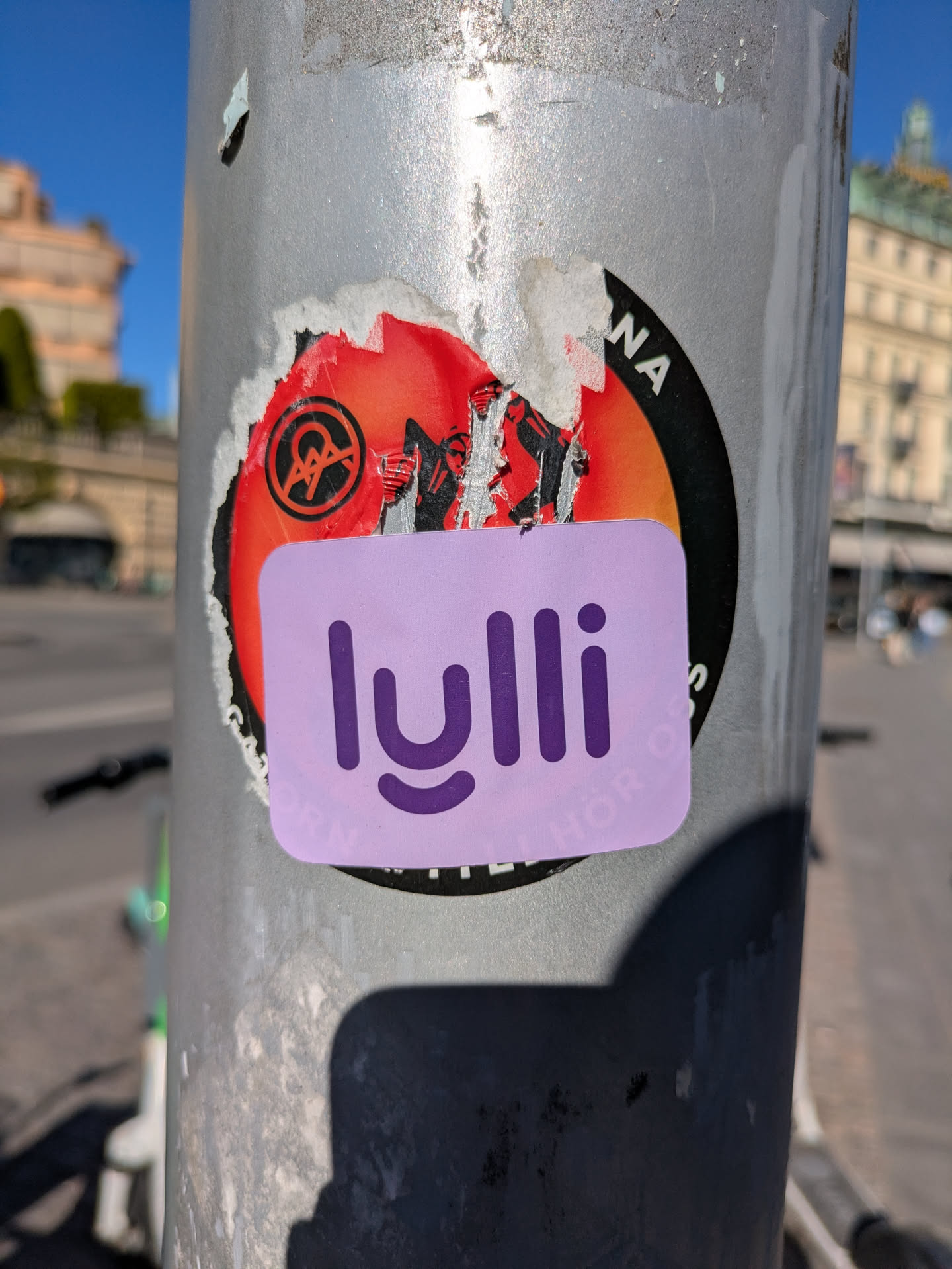A lavender rectangular sticker with rounded edges features the word "lulli" in a stylized purple font. The sticker is adhered to a light pole, partially covering another sticker that is red, black, and white. The text under the lulli logo says "PINK, BUT ANCHOR DOWN".
