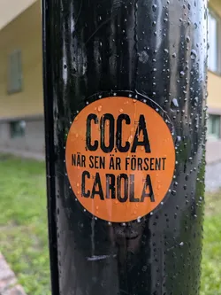 The sticker is circular with a vibrant orange background. It features bold, black, distressed lettering. The top line reads 'COCA' in large, capitalized letters. Below this, in smaller text, it says 'NÄR SEN ÄR FÖRSENT'. The bottom line prominently displays 'CAROLA' in large, capitalized letters. The edge of the sticker is decorated with a pattern of small white stars. The sticker appears to be adhered to a wet, black cylindrical object, with water droplets visible on both the sticker and the surface it's attached to.