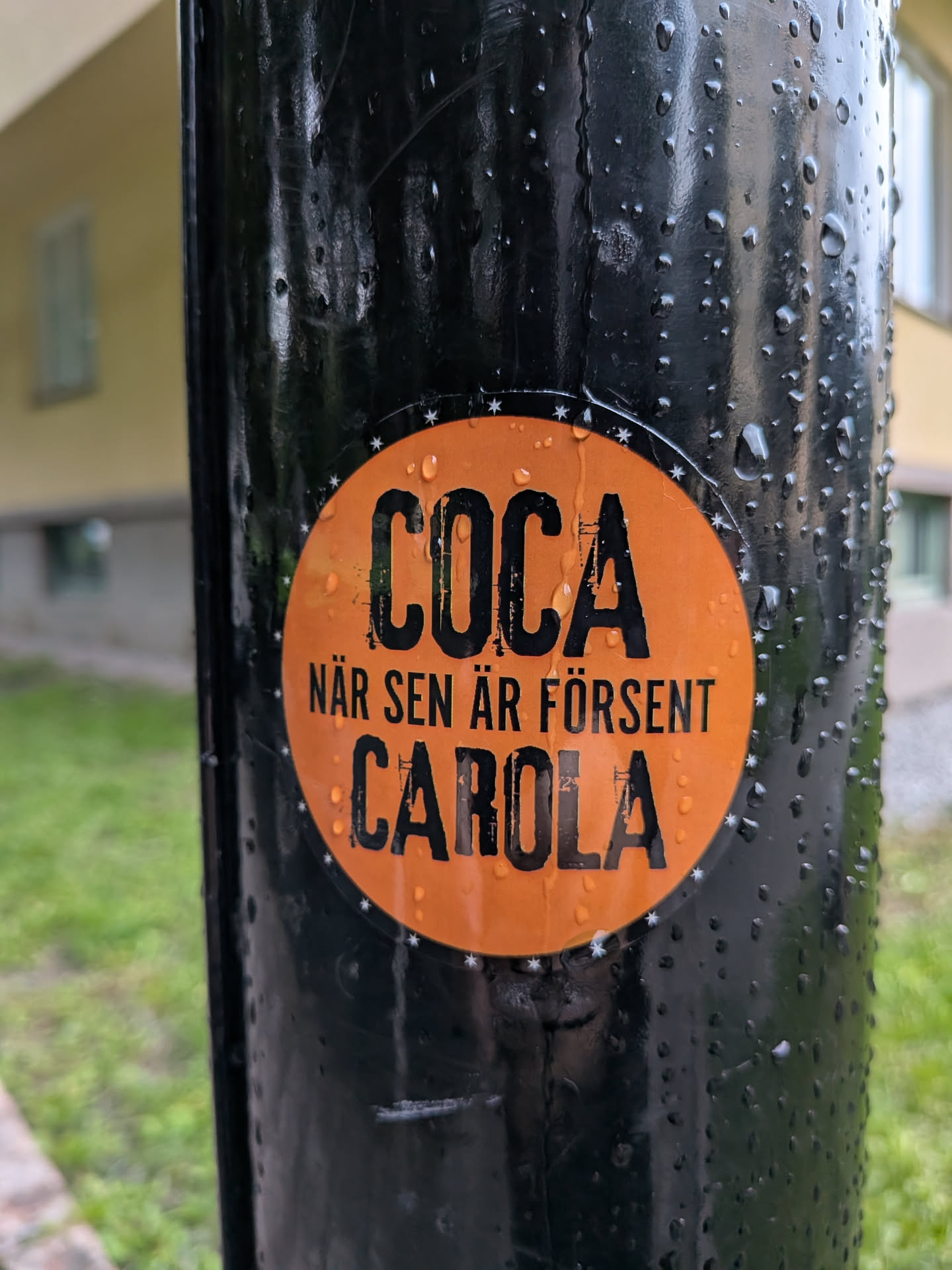 The sticker is circular with a vibrant orange background. It features bold, black, distressed lettering. The top line reads 'COCA' in large, capitalized letters. Below this, in smaller text, it says 'NÄR SEN ÄR FÖRSENT'. The bottom line prominently displays 'CAROLA' in large, capitalized letters. The edge of the sticker is decorated with a pattern of small white stars. The sticker appears to be adhered to a wet, black cylindrical object, with water droplets visible on both the sticker and the surface it's attached to.