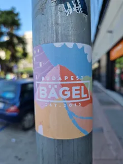 A colorful sticker featuring a stylized illustration of Budapest with the words "BUDAPEST B'AGEL EST. 2013." The design has a vintage, illustrated feel with pastel colors.