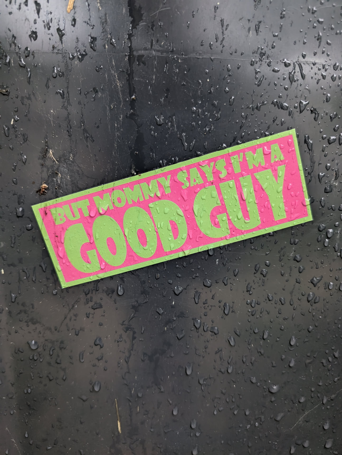 A rectangular sticker with a bright pink background and neon green text. The text is in a bold, blocky font. The top line reads 'BUT MOMMY SAYS I'M A' and the bottom, larger line reads 'GOOD GUY'. The sticker is slightly angled and appears to be adhered to a dark, textured surface covered in water droplets.