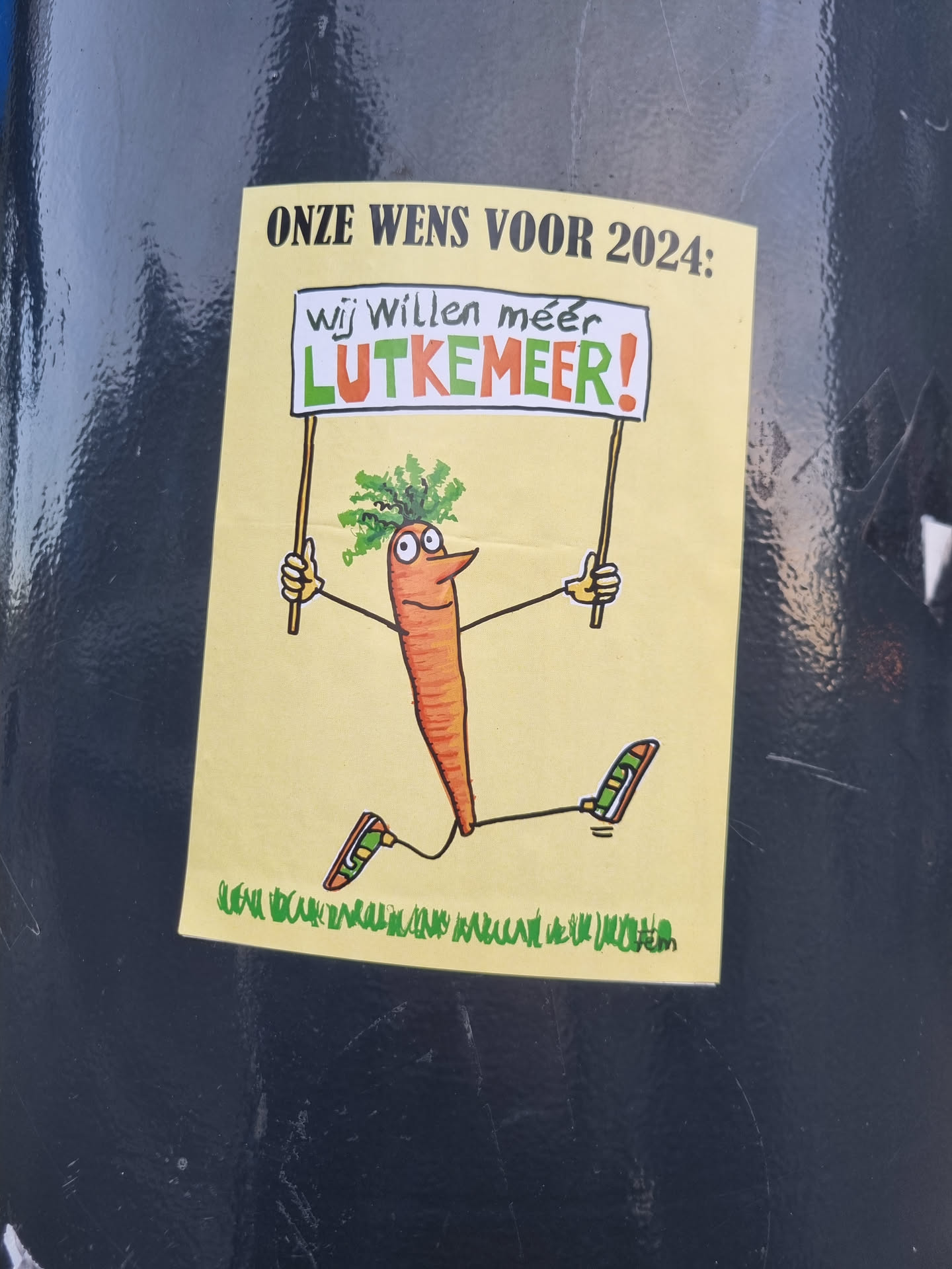 A sticker featuring a cartoon carrot character holding a sign that says "We want more Lutkemeer!" in Dutch. The carrot is depicted as running, and the text at the bottom seems to be a smaller tagline or additional information.