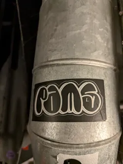 Street sticker A rectangular sticker placed horizontally on a galvanized metal duct. The sticker features the word 'rams' rendered in a classic graffiti throw-up or bubble letter style with thick black fill and concentric white outlines against a solid black background. Small white characters resembling 'L. Y' are nested inside the final letter 's'.