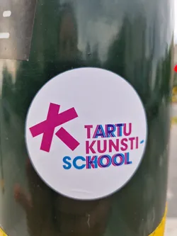 Round white sticker with a pink stylized letter "K" and the text "TARTU KUNSTI-SCOOL" in a colorful, slightly distorted font. The sticker is affixed to a dark green pole.