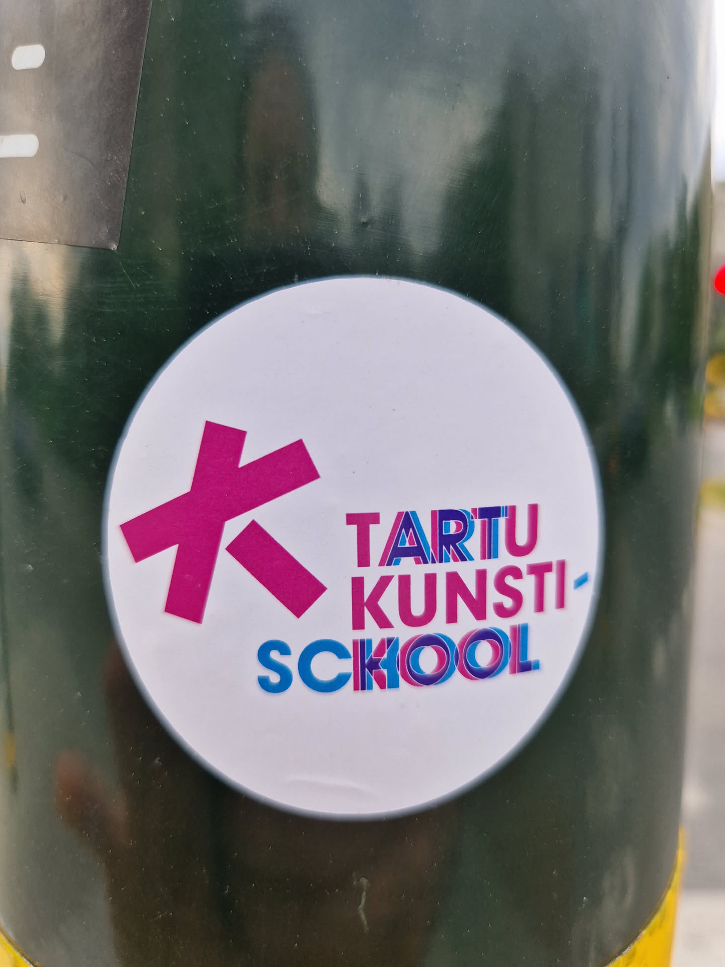 Round white sticker with a pink stylized letter "K" and the text "TARTU KUNSTI-SCOOL" in a colorful, slightly distorted font. The sticker is affixed to a dark green pole.