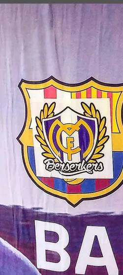 Street sticker The sticker features a custom football crest, prominently displayed on what appears to be a larger, faded FC Barcelona-themed background. The main crest is a fusion of two iconic Spanish football clubs. The outer shield shape and its upper half incorporate the vertical red and yellow stripes of the Catalan flag, characteristic of FC Barcelona's emblem, along with blue and red stripes in the lower half. Overlaid on this is a smaller, central shield in purple with a golden 'MCF' monogram, reminiscent of Real Madrid's crest, surrounded by a golden laurel wreath. Below this central design, the word 'Berserkers' is written in a stylized, flowing white script with a black outline. The sticker itself has clear, slightly raised edges, indicating it's a physical adhesive item placed on another surface.