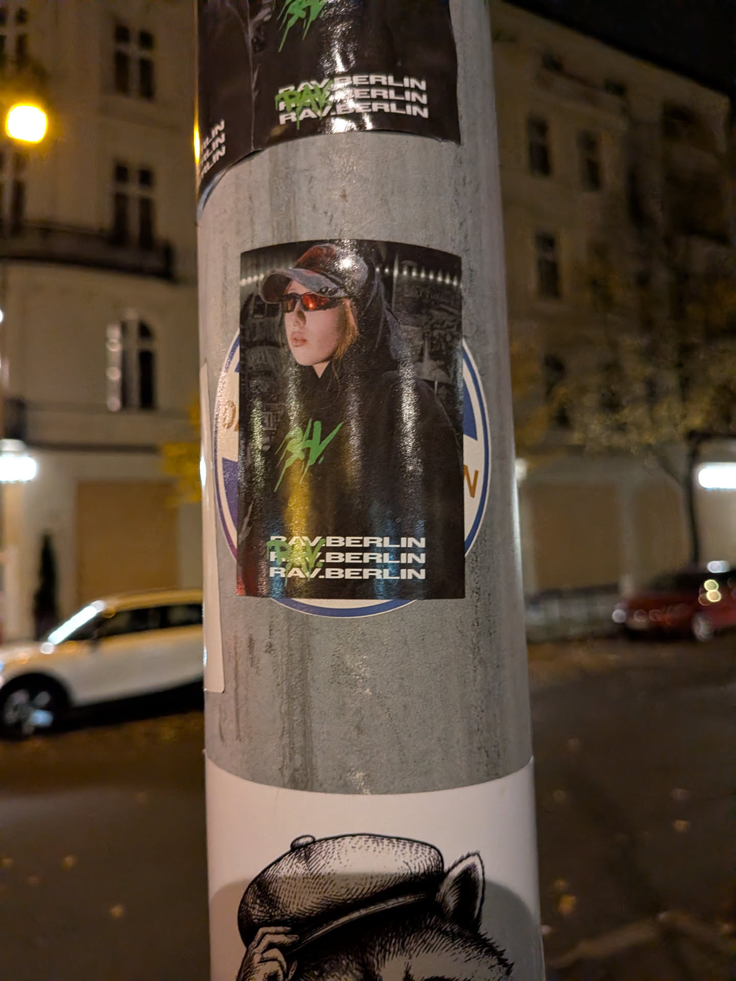 A vertical rectangular sticker depicting a person wearing a cap, reflective wraparound sunglasses, and a black hoodie featuring a neon green spiky graphic on the chest. Across the bottom of the sticker, the text 'RAV.BERLIN' is printed three times in bold white uppercase font, overlaid on the left by the same green emblem.