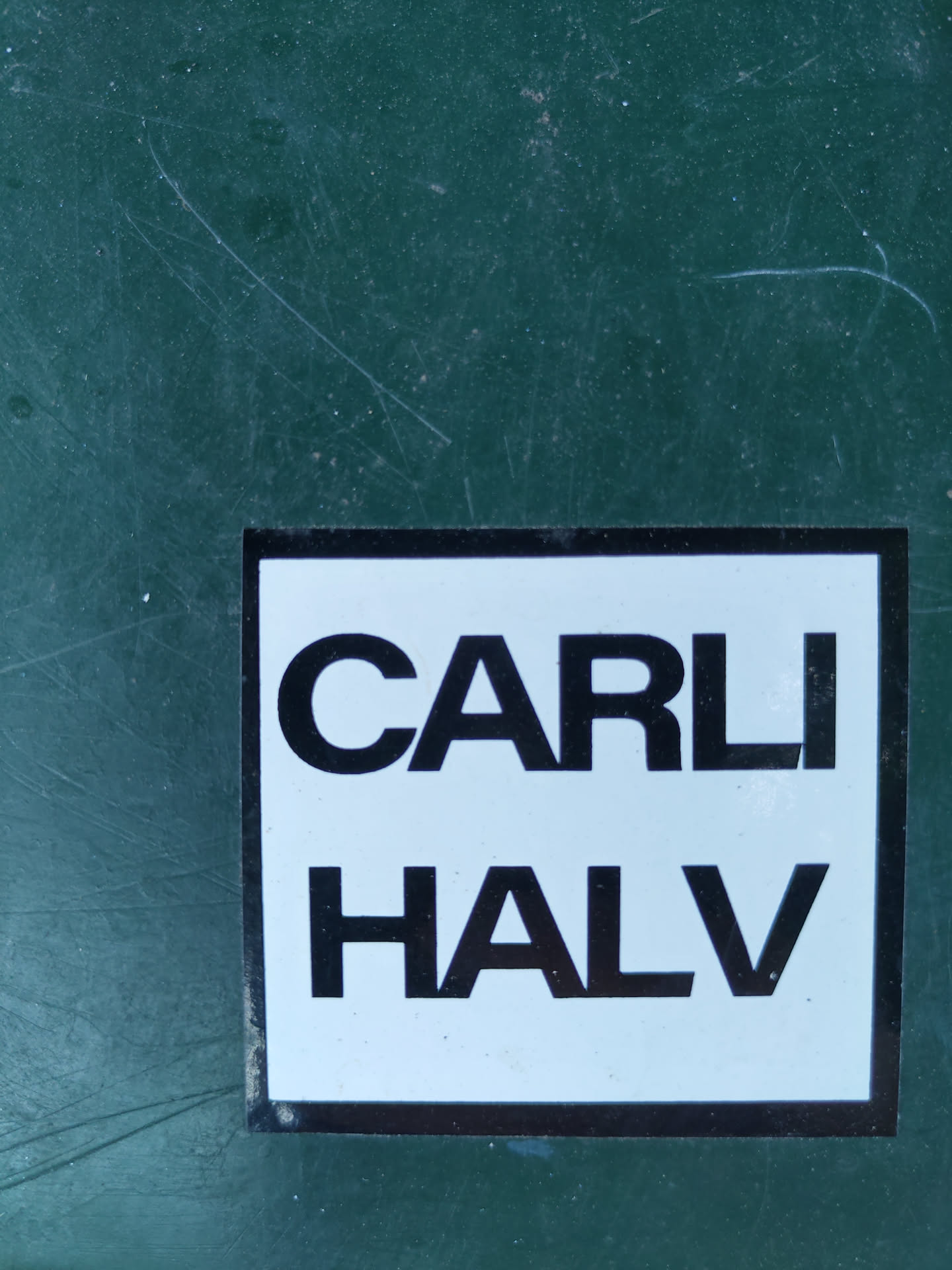 A white sticker with black text that says "CARLI HALV" on a dark green surface.
