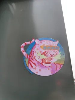 A round sticker featuring a stylized pink tiger with yellow mustache-like markings. The tiger has pink polka dots and is set against a blue and yellow retro background. Text in Asian characters is visible across the tiger's face. The bottom of the sticker shows the website johnyee.com