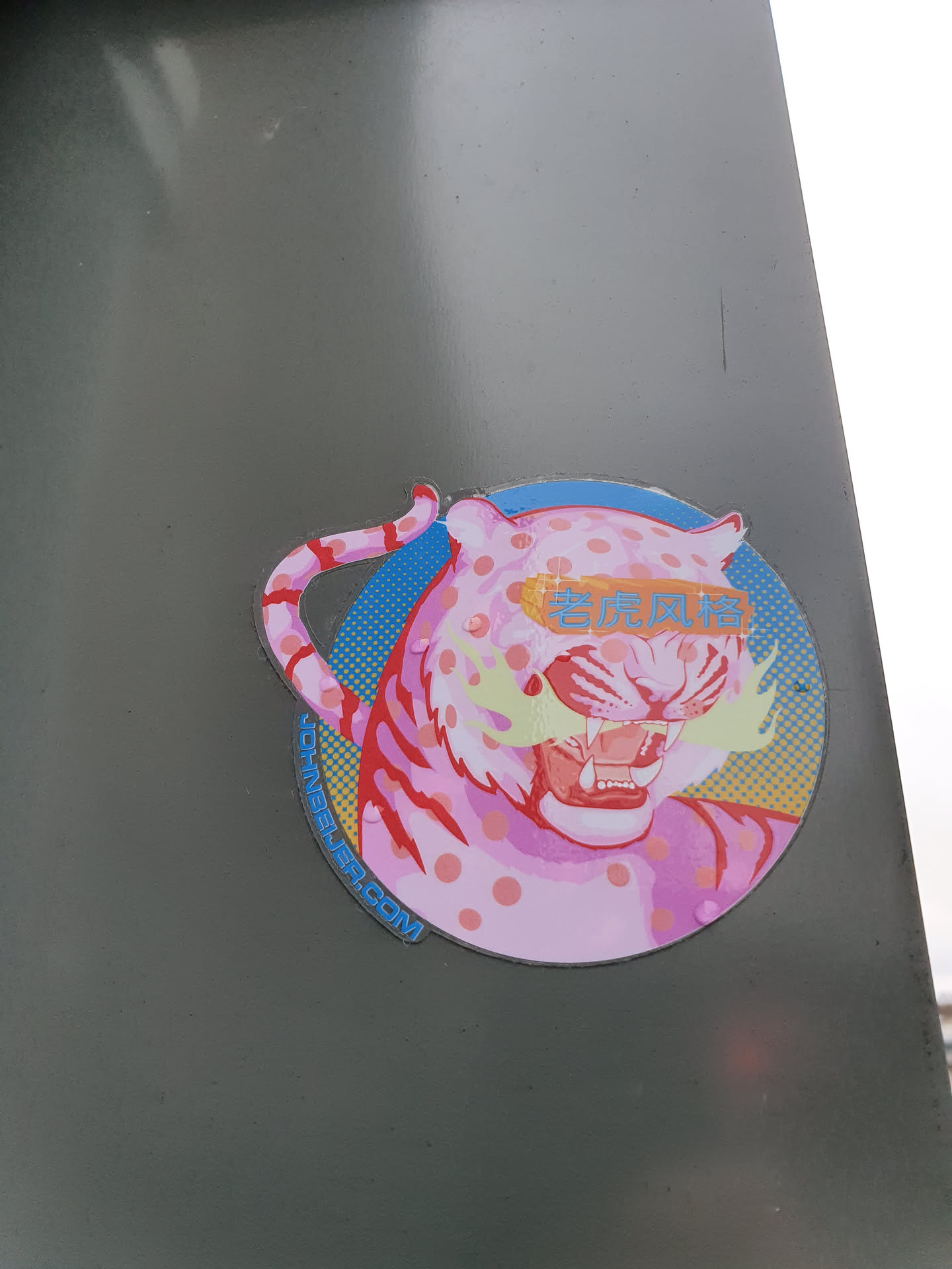 A round sticker featuring a stylized pink tiger with yellow mustache-like markings. The tiger has pink polka dots and is set against a blue and yellow retro background. Text in Asian characters is visible across the tiger's face. The bottom of the sticker shows the website johnyee.com