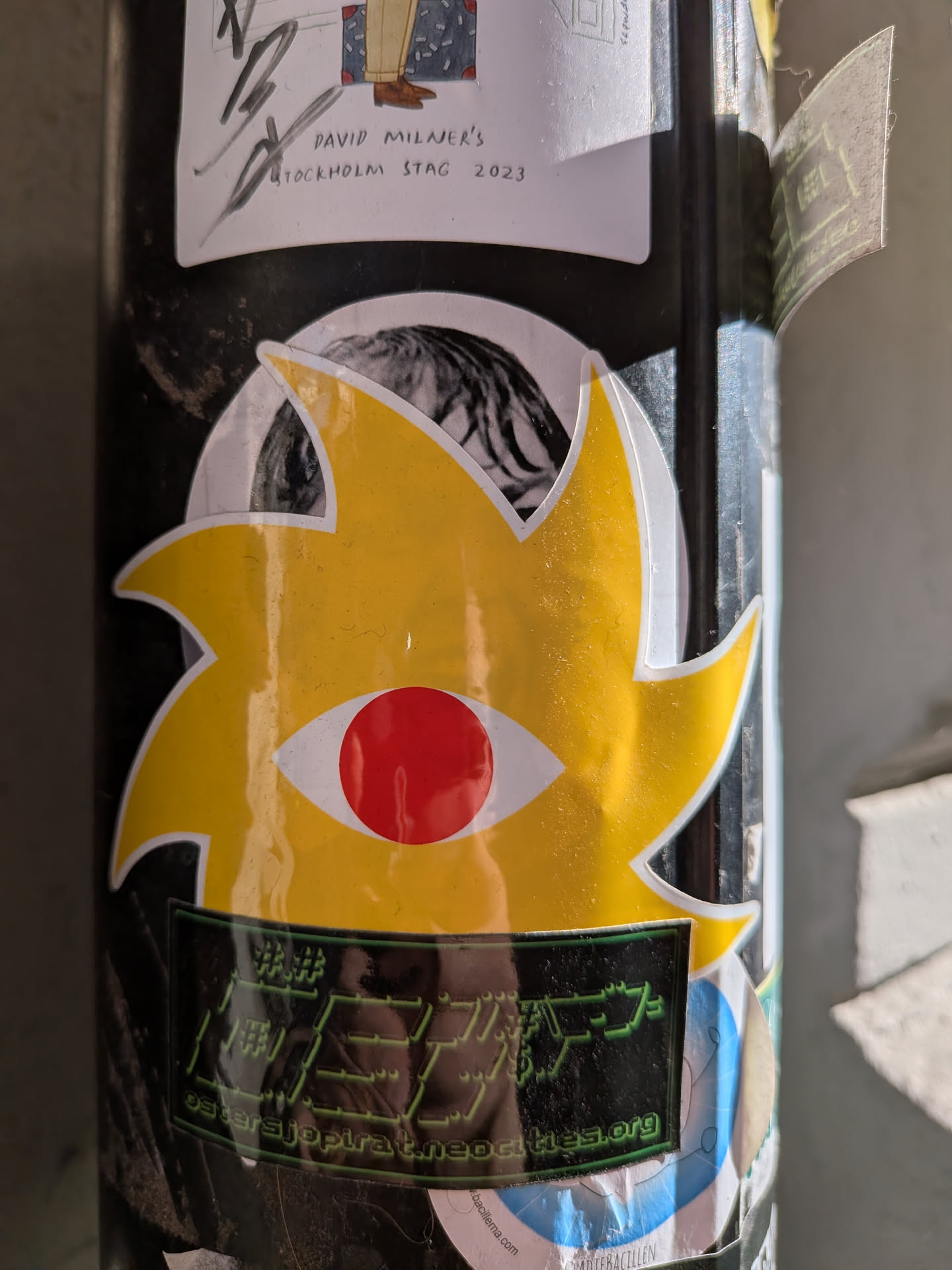 The central sticker is a striking, abstract design predominantly in bright yellow with a clean white outline. Its shape is reminiscent of a spiky star or a stylized sun, with multiple pointed protrusions. At its center is a prominent eye motif, featuring a white sclera and a bold, circular red pupil or iris. Below this yellow eye, integrated into the sticker's lower portion, is a rectangular black area with a glowing green outline. This black section contains pixelated green text, which is partially obscured and stylized, and a clearly readable URL at the bottom.