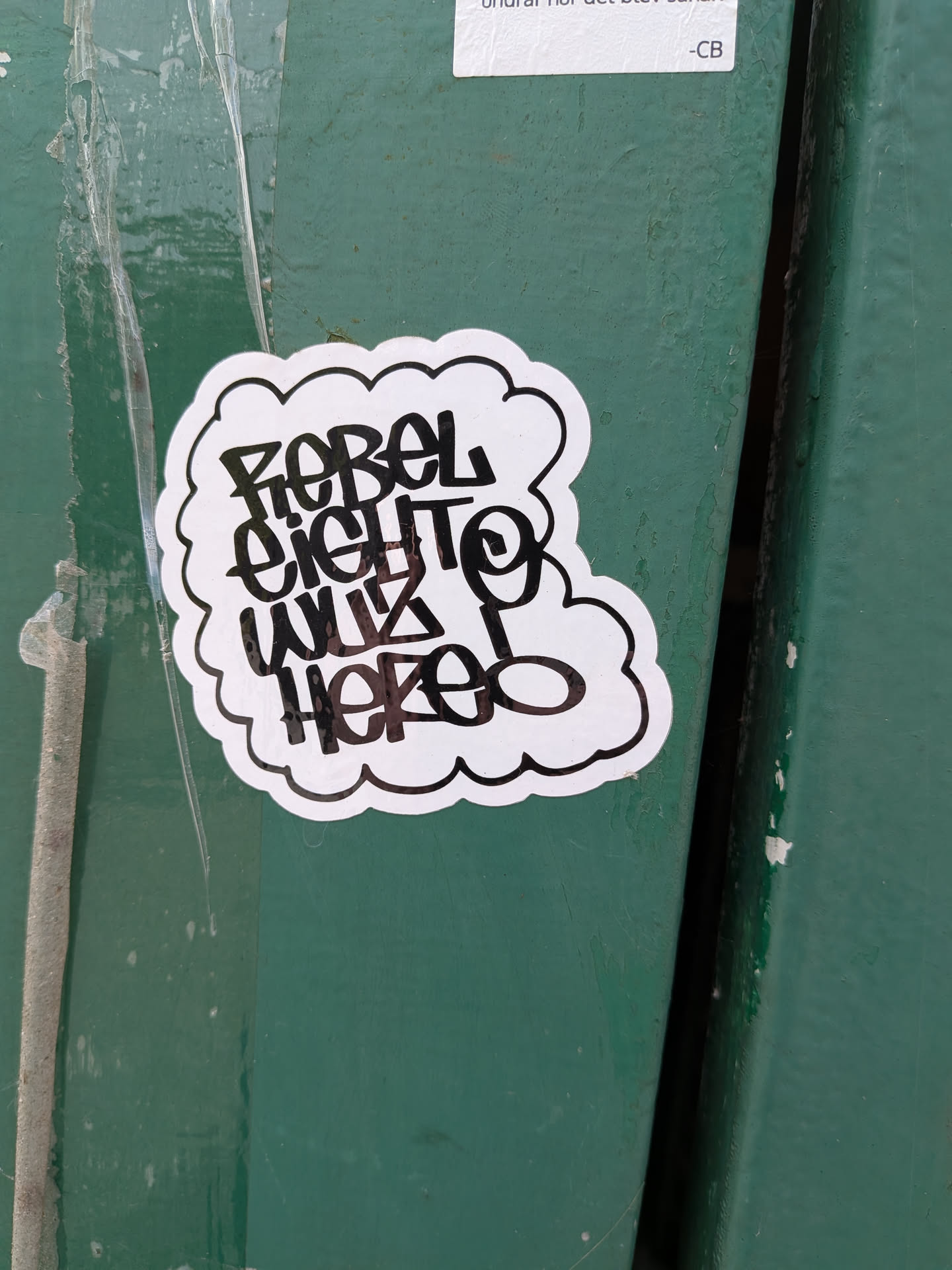 A die-cut, cloud-shaped sticker with a white background and a thin black border. The center features stylized, hand-written graffiti text in black ink that reads 'REBEL EIGHT WUZ HERE!'. The sticker is affixed to a textured, green-painted surface.
