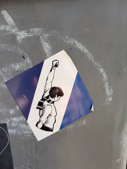 The sticker shows a person with a raised fist, wearing a scarf. The sticker's background is split diagonally, with one section blue and the other white. It's stuck to a gray surface.