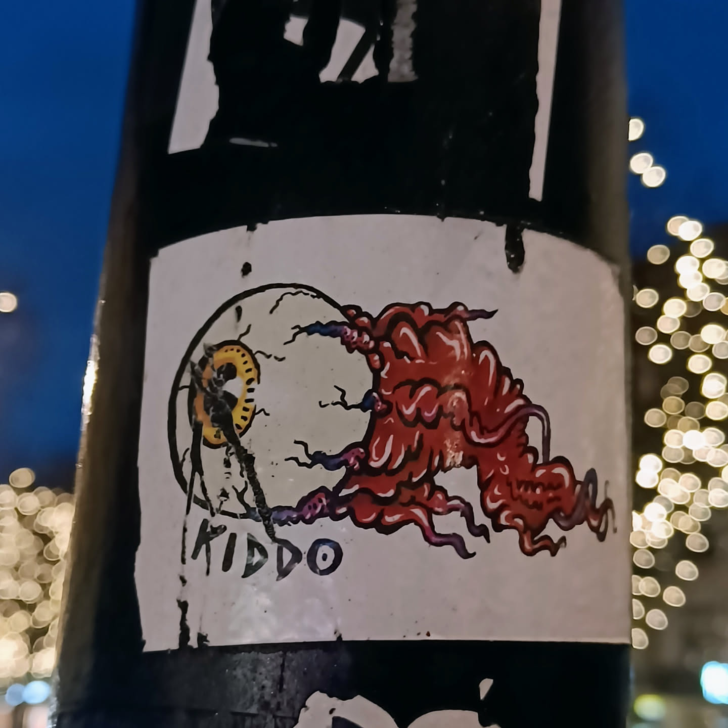 A sticker featuring a creepy eyeball with a red, fleshy appendage emerging from it. The style is somewhat reminiscent of horror or fantasy art. The sticker is slightly damaged, with a tear near the bottom.