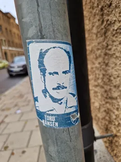 A blue and white sticker featuring a portrait of Torø Mansæn. The sticker is affixed to a metal pole on a city street.