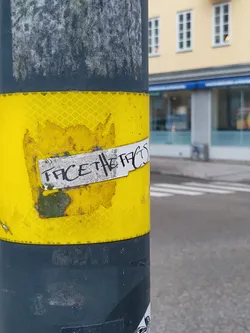 A worn sticker affixed to a yellow and gray pole, boldly proclaiming the message "Face the facts". The sticker shows signs of weathering and damage, adding to its street aesthetic.