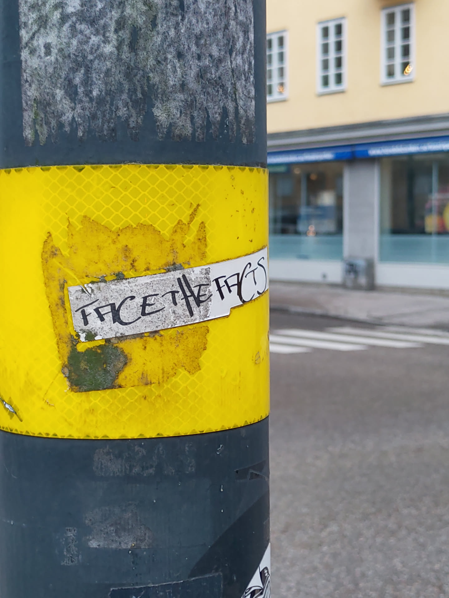 A worn sticker affixed to a yellow and gray pole, boldly proclaiming the message "Face the facts". The sticker shows signs of weathering and damage, adding to its street aesthetic.